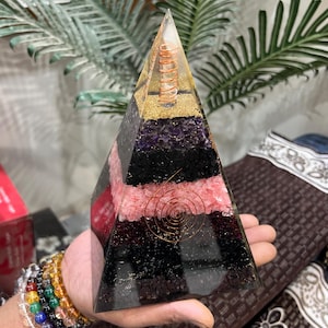 Puede incluir: Una pir&aacute;mide de cristal transparente con capas de amatista, cuarzo rosa, turmalina negra y purpurina dorada. Una bobina de alambre de cobre est&aacute; incrustada en el centro de la pir&aacute;mide.
