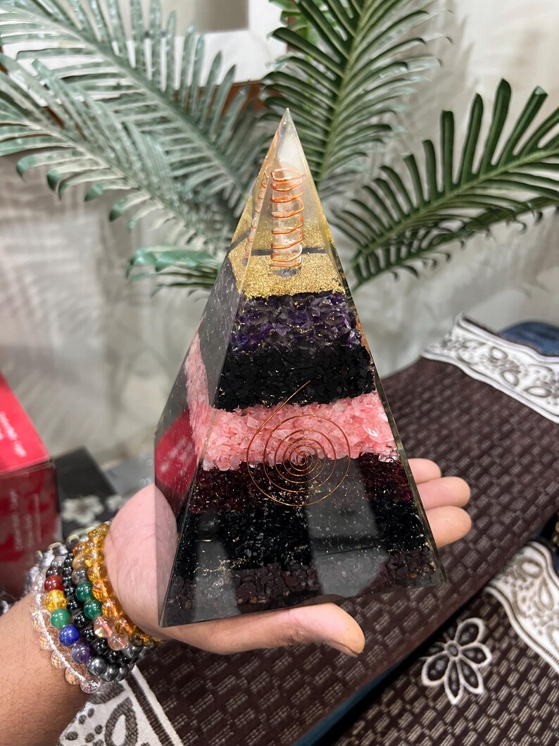 Puede incluir: Una pir&aacute;mide de cristal transparente con capas de amatista, cuarzo rosa, turmalina negra y purpurina dorada. Una bobina de alambre de cobre est&aacute; incrustada en el centro de la pir&aacute;mide.