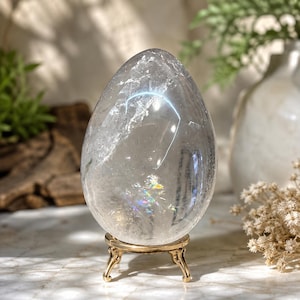 May include: A clear, egg-shaped quartz crystal with visible internal fractures and rainbow light refractions. The crystal is polished to a smooth finish and rests securely on a small, three-legged, polished gold-tone metal display stand.