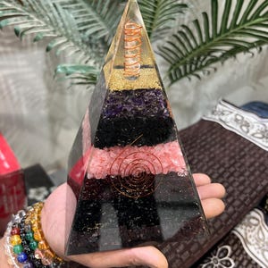 Puede incluir: Una pir&aacute;mide transparente de resina con capas de cristales de amatista, turmalina negra, cuarzo rosa y cuarzo claro. Una bobina de alambre de cobre est&aacute; incrustada en el centro de la pir&aacute;mide.