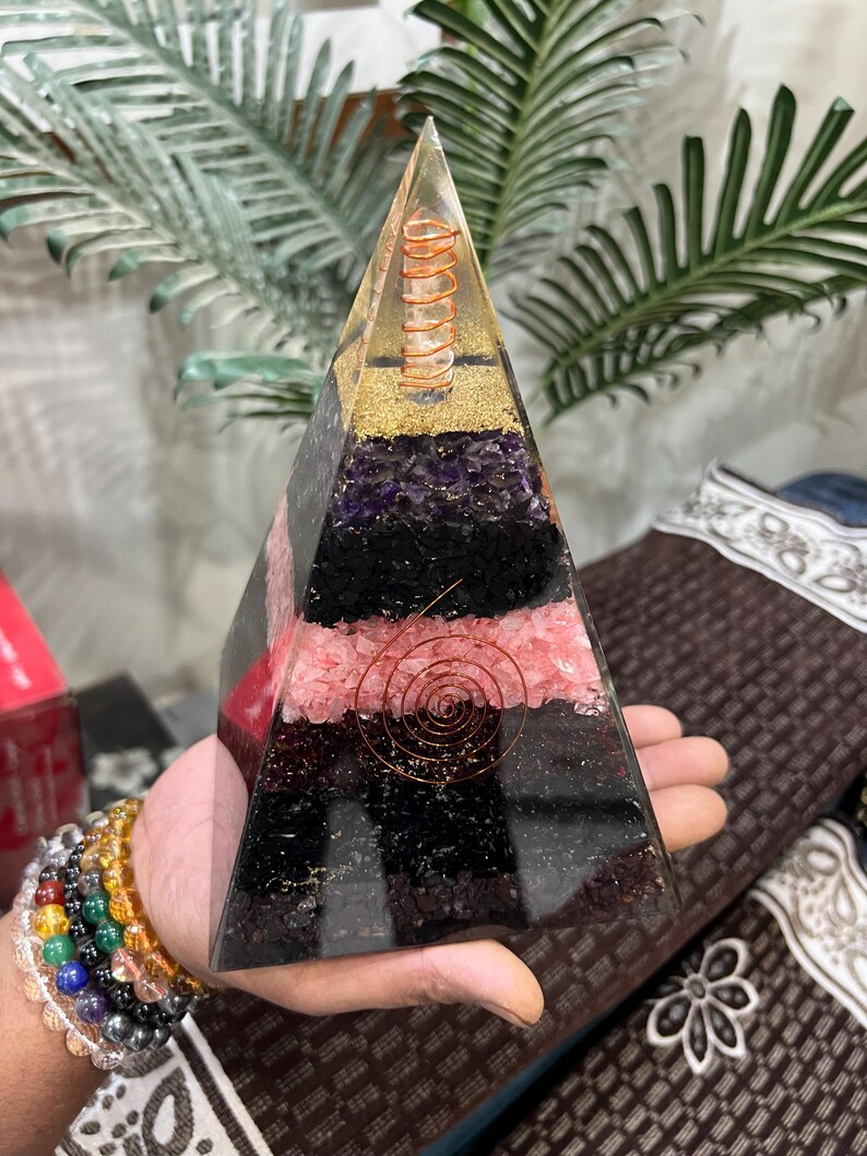 Puede incluir: Una pir&aacute;mide transparente de resina con capas de cristales de amatista, turmalina negra, cuarzo rosa y cuarzo claro. Una bobina de alambre de cobre est&aacute; incrustada en el centro de la pir&aacute;mide.