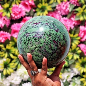 Könnte beinhalten: Eine grüne und rosafarbene Edelsteinkugel, poliert zu einem glatten Finish. Die Kugel wird in einer Hand gehalten und zeigt ihre komplizierten Muster und Farben.