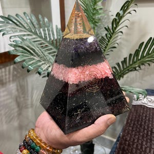 Puede incluir: Una pir&aacute;mide de cristal en capas con una espiral de alambre de cobre en su interior. La pir&aacute;mide est&aacute; hecha de cristales de amatista, cuarzo rosa y obsidiana negra. La pir&aacute;mide mide aproximadamente 15 cm de alto.