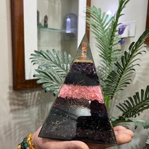 Puede incluir: Una pir&aacute;mide transparente hecha de cristales en capas, incluyendo amatista, turmalina negra, cuarzo rosa y cuarzo claro. Una bobina de alambre de cobre est&aacute; incrustada en la capa de cuarzo rosa. La pir&aacute;mide est&aacute; siendo sostenida en una mano.