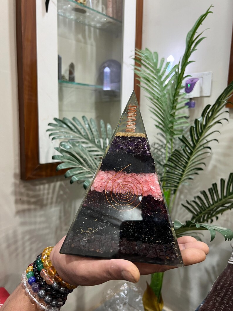 Puede incluir: Una pir&aacute;mide transparente hecha de cristales en capas, incluyendo amatista, turmalina negra, cuarzo rosa y cuarzo claro. Una bobina de alambre de cobre est&aacute; incrustada en la capa de cuarzo rosa. La pir&aacute;mide est&aacute; siendo sostenida en una mano.