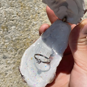 May include: A silver ring with a small, round, clear gemstone is displayed inside an open oyster shell. The shell is white with iridescent glitter. The ring is held in a person's hand against a textured, light-colored background.