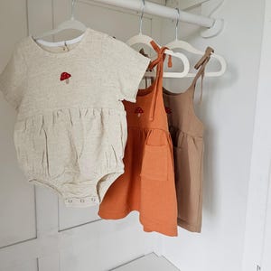 Pode incluir: Três roupas de bebê penduradas em cabides brancos. As roupas incluem um macacão bege com uma aplicação de cogumelo, um vestido laranja com um bolso e um vestido marrom. Todos são feitos de um tecido texturizado.