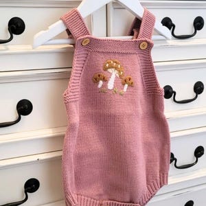 Pode incluir: Um macacão de malha rosa para bebê com um design de bordado de cogumelos. O macacão tem um fecho de botão na parte inferior.