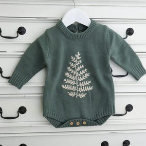 Puede incluir: Un pelele de punto verde con un diseño de árbol bordado en blanco. El pelele tiene un cuello redondo y mangas largas.