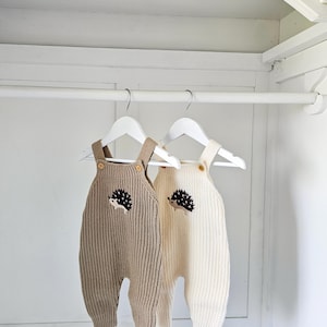 May include: Two knitted baby dungarees with hedgehog appliques hanging on hangers in a white wardrobe. One dungaree is beige and the other is white.
