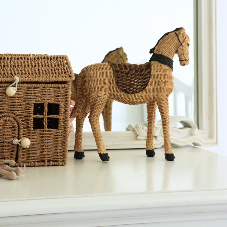 Puede incluir: Una figura de caballo de mimbre tejido con una silla de montar marr&oacute;n oscuro y cascos negros se encuentra sobre una superficie blanca. A la izquierda hay una cesta a juego con forma de casa de mimbre con tapa con bisagras y una peque&ntilde;a ventana. Un espejo refleja el caballo.