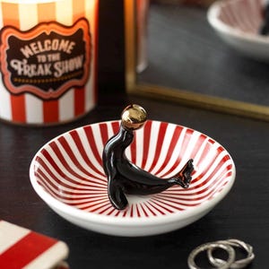 May include: A small, white ceramic dish with a red and white striped pattern, holding a black seal figurine with a gold-coloured ball on its head. The dish is on a dark surface, with a candle and mirror in the background.