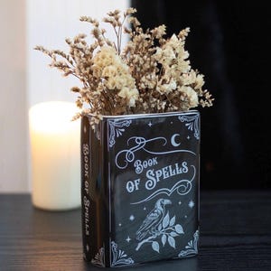 May include: A black ceramic vase in the shape of a book, inscribed with "Book of Spells" in white lettering. The vase has a raven illustration and contains dried, cream-coloured flowers. A lit candle is in the background.