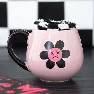 May include: A pink ceramic mug with a black handle and a black flower design with a sad face. The mug is filled with a black and white checkered item. The mug is on a dark surface.