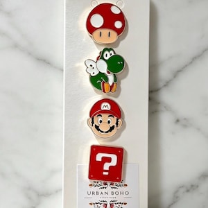 May include: A set of four enamel pins featuring a red and white mushroom, a green dinosaur with wings, a character with a red hat and moustache, and a red square block with a white question mark, all displayed on a white card labelled Urban Boho.