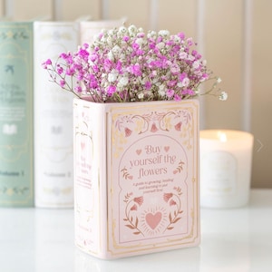 May include: A pink ceramic vase in the shape of a book, holding pink and white flowers. The vase features gold detailing and the words "Buy yourself the flowers". Other books and a candle are in the background.
