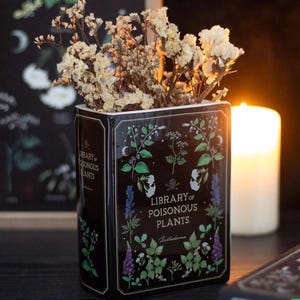 May include: A black ceramic vase with the text "Library of Poisonous Plants" and botanical illustrations. The vase is filled with dried, cream-coloured flowers. A lit candle is in the background.