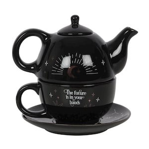 May include: Black ceramic teapot and teacup set with a saucer. The teapot features a celestial design with a crescent moon and sunburst. The teacup has the text "The future is in your hands".