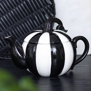 May include: A black and white striped ceramic teapot shaped like a pumpkin. The spout, handle, and lid handle are black. The teapot is decorated with vertical stripes and sits on a dark surface.