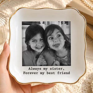 May include: A square white ceramic trinket dish with scalloped edges and a gold-painted rim. The center features a black and white photograph of two young girls with the printed text: Always my sister, Forever my best friend.