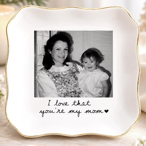 May include: A square white ceramic trinket dish with scalloped edges and a gold-painted rim. The center features a black and white photograph of a woman and a child, with the printed text "I love that you're my mom" followed by a small heart.