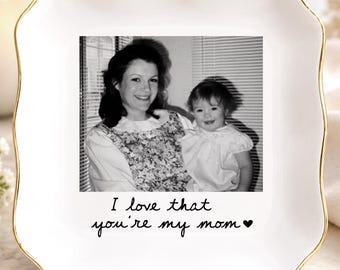 May include: A square white ceramic trinket dish with scalloped edges and a gold-painted rim. The center features a black and white photograph of a woman and a child, with the printed text "I love that you're my mom" followed by a small heart.