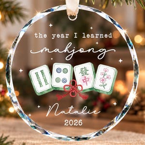 以下が含まれることがあります： 「the year I learned mahjong」、「Natalie」、「2026」という文字が入った、丸い透明なガラス製のクリスマスオーナメント。4枚の麻雀牌と赤い装飾結びが描かれており、白いリボンで吊り下げられています。