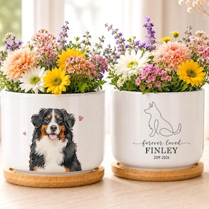 May include: Two white ceramic cylindrical planters with bamboo saucers, each filled with colorful flowers. One planter features a Bernese Mountain Dog illustration with small pink hearts. The other shows a line-art dog with the text forever loved FINLEY 2019-2026.