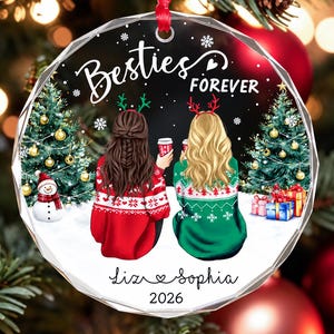 May include: A round, faceted glass Christmas ornament featuring an illustration of two people from behind, wearing holiday sweaters and reindeer headbands. The text reads Besties FOREVER, Liz, Sophia, and 2026, with snowy trees and gifts.