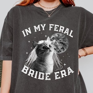 May include: A dark gray t-shirt with the text "In My Feral Bride Era" in white. A black and white image of a cat wearing a veil is in the center of the shirt.