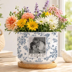 May include: A white ceramic planter with a blue floral pattern, featuring a black and white photograph of two people and the text "Be the things you loved the most about her" with a heart icon. The planter sits on a round wooden base.