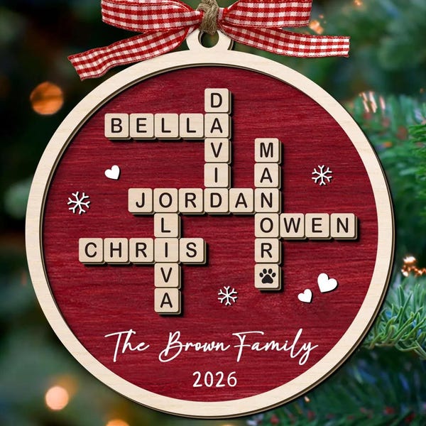 Puede incluir: Un adorno navideño redondo de madera con fondo de veta de madera roja que presenta un diseño de crucigrama con los nombres Bella, David, Jordan, Olivia, Chris y Owen. Incluye una cinta de cuadros rojos y blancos y el texto The Brown Family 2026.