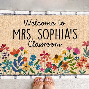 Może przedstawiać: Prostokątna wycieraczka z czarnym napisem "Welcome to MRS. SOPHIA'S Classroom". Mata ma beżowe tło z kwiatową obw&oacute;dką z kolorowymi kwiatami. Obramowanie jest obramowane czarno-białym wzorem w kratę.