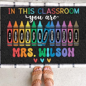 May include: A black and white welcome mat with the text "In this classroom you are" and a row of colorful crayons with words like "kind", "creators", "special", "important", "trusted", "smart", "unique", "respected", "loved", "amazing", "valued", and "good" written on them. The mat also has the text "Mrs. Wilson" in colorful letters.
