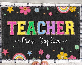 May include: A black welcome mat with the word "TEACHER" in colorful letters with gold glitter. The mat also has the text "Mrs. Sophia" and a rainbow, stars, and flowers design.