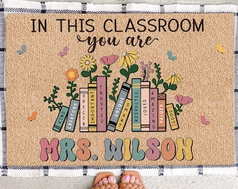 May include: A welcome mat with a brown burlap background and the text "IN THIS CLASSROOM you are" in brown lettering.  The text is surrounded by colorful flowers and books with words like "KIND", "CREATORS", "TRUSTED", "IMPORTANT", "UNIQUE", "SMART", "RESPECTED", "LOVED", "AMAZING", "VALUED", and "GOOD".  The text "MRS. WILSON" is in colorful, stylized lettering at the bottom of the mat.