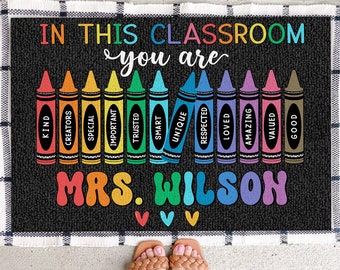May include: A black and white welcome mat with the text "In this classroom you are" and a row of colorful crayons with words like "kind", "creators", "special", "important", "trusted", "smart", "unique", "respected", "loved", "amazing", "valued", and "good" written on them. The mat also has the text "Mrs. Wilson" in colorful letters.