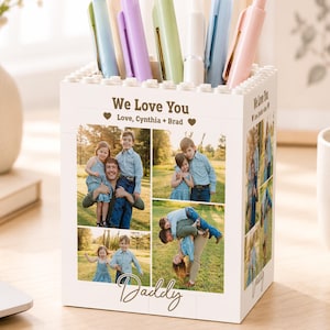 May include: A white pen holder with a brick-like design, personalized with family photos and the text "We Love You" and "Daddy." The holder contains various colored pens. A thoughtful gift for a loved one.