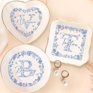 May include: Three white ceramic trinket dishes with blue floral designs and gold rims. One is heart-shaped, one square, and one round. Each dish features a different initial and name in blue script: "Victoria", "Taylor", and "Brittany". Two gold earrings with pearl drops are also visible.