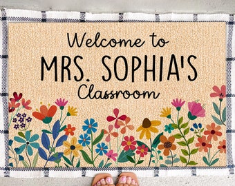 May include: A rectangular doormat with the text "Welcome to MRS. SOPHIA'S Classroom" in black. The mat has a beige background with a floral border featuring colorful flowers. The border is framed by a black and white checkered pattern.