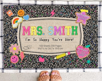 May include: A doormat with a notebook design, featuring the text "MRS. SMITH I'm So Happy You're Here!". It includes colorful embroidered patches of a flower, smiley faces, a rainbow, and a pencil. The mat is 9.75 inches by 7.5 inches.