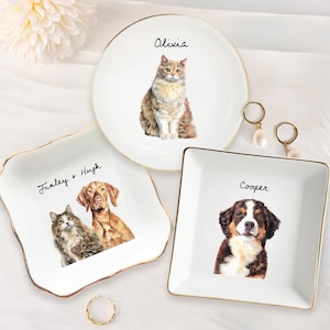 May include: Three ceramic trinket dishes with gold trim. The dishes feature illustrations of pets and their names. The first dish is square and features a cat and dog with the names Finley and Hugh. The second dish is round and features a cat named Olivia. The third dish is square and features a dog named Cooper.