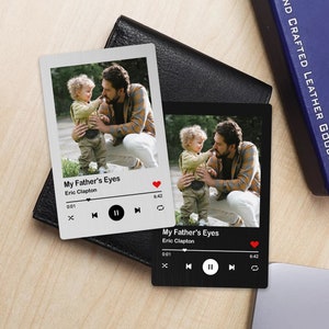 May include: Two silver and black metal music player coasters with a photo of a father and son. The coasters feature a play button, pause button, and a skip button. The text on the coasters reads "My Father's Eyes" and "Eric Clapton".