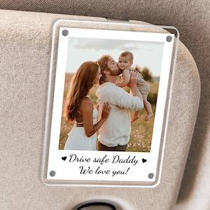 May include: A clear acrylic car visor photo holder with a family photo. The photo shows a family of three in a field. The text on the holder reads "Drive safe Daddy, We love you!" in black script. The holder is attached to a car visor.