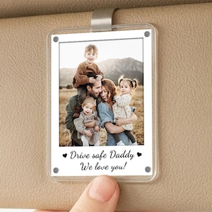 May include: A clear, rectangular car visor photo holder with a family photo. The photo features a family of five, with the text "Drive safe Daddy We love you!" in black. The holder is attached to a beige car visor.