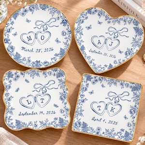 May include: Four white ceramic trinket dishes with gold-painted rims, featuring blue floral patterns, butterflies, and a central locket illustration. Each dish displays unique initials and dates: M O March 28, 2026; A J September 25, 2026; L T September 19, 2026; and T K April 4, 2025.