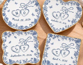 May include: Four white ceramic trinket dishes with gold-painted rims, featuring blue floral patterns, butterflies, and a central locket illustration. Each dish displays unique initials and dates: M O March 28, 2026; A J September 25, 2026; L T September 19, 2026; and T K April 4, 2025.