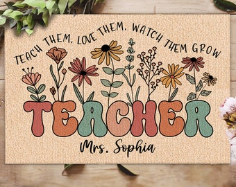 May include: A tan doormat with the word "TEACHER" in colorful, retro-style letters. The text "Teach them, love them, watch them grow" is above the word "TEACHER" and the text "Mrs. Sophia" is below the word "TEACHER". The background is a tan burlap-like material with a floral design.