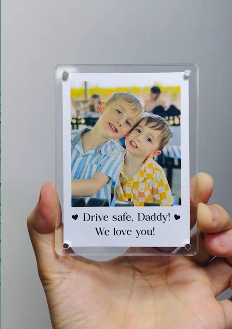 May include: A clear acrylic photo frame holding a printed photograph of two young boys smiling. The photo includes the text "Drive safe, Daddy! We love you!" in black font, with a heart symbol on each side.