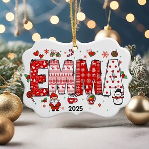 May include: A white ceramic Christmas ornament with the name "EMMA" in red, festive lettering. The letters are decorated with holiday patterns and symbols, including snowflakes, Santa, and a snowman. The year "2025" is also displayed.